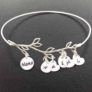 May include: Silver bangle bracelet with a delicate branch design and hanging charms. The charms are round and heart-shaped with the letters "Nana", "R", "A", "L", "M", and "L" engraved on them.