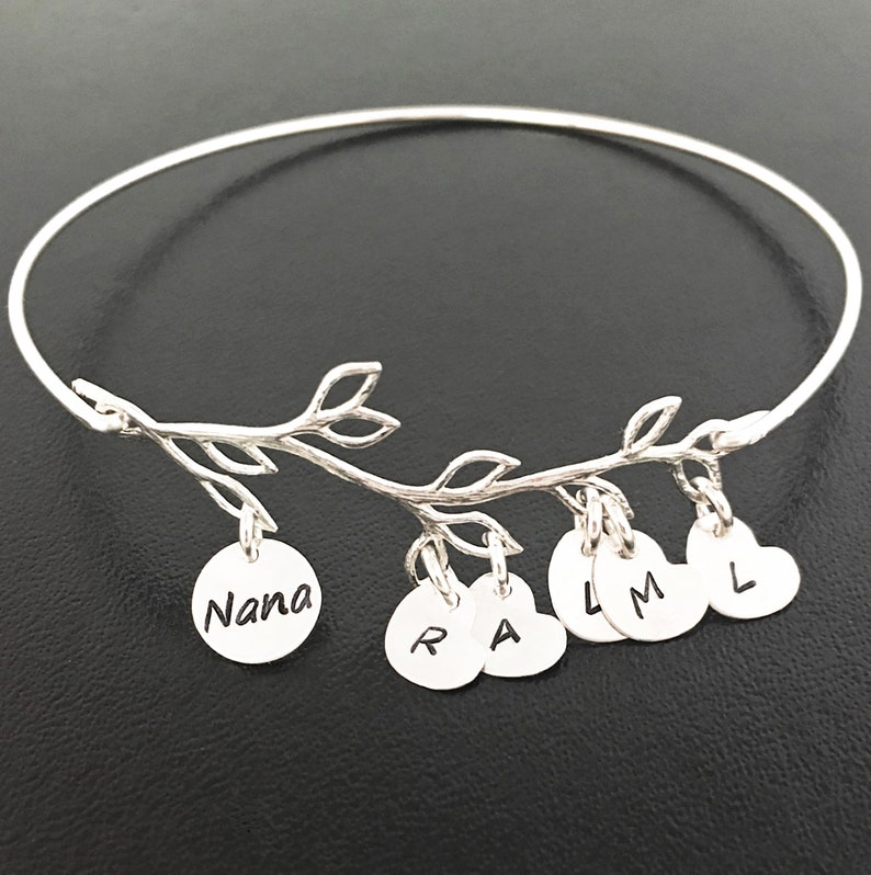 May include: Silver bangle bracelet with a delicate branch design and hanging charms. The charms are round and heart-shaped with the letters "Nana", "R", "A", "L", "M", and "L" engraved on them.