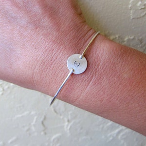 May include: A silver bangle bracelet with a round charm featuring the letter "E" engraved on it.