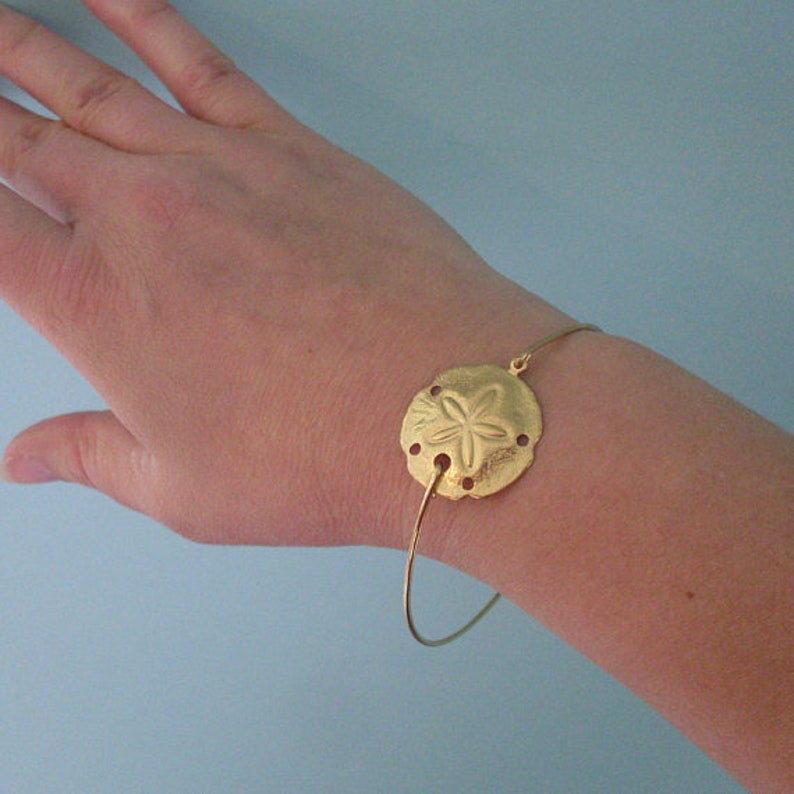 May include: A gold-toned bangle bracelet with a sand dollar charm. The charm is detailed with a five-pointed star in the center.