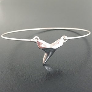 May include: A silver shark tooth charm hangs from a silver bangle bracelet. The bracelet is open and ready to be worn.