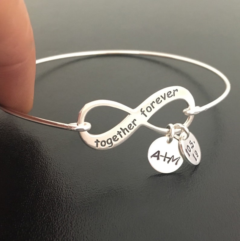 May include: Silver infinity bracelet with the words "together forever" engraved on the infinity symbol. The bracelet has two charms, one with the initials "A+M" and the other with the date "10.5.19".