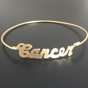 May include: Gold bangle bracelet with the word "Cancer" written in a cursive font.