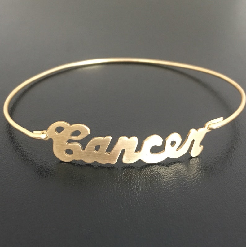 May include: Gold bangle bracelet with the word "Cancer" written in a cursive font.