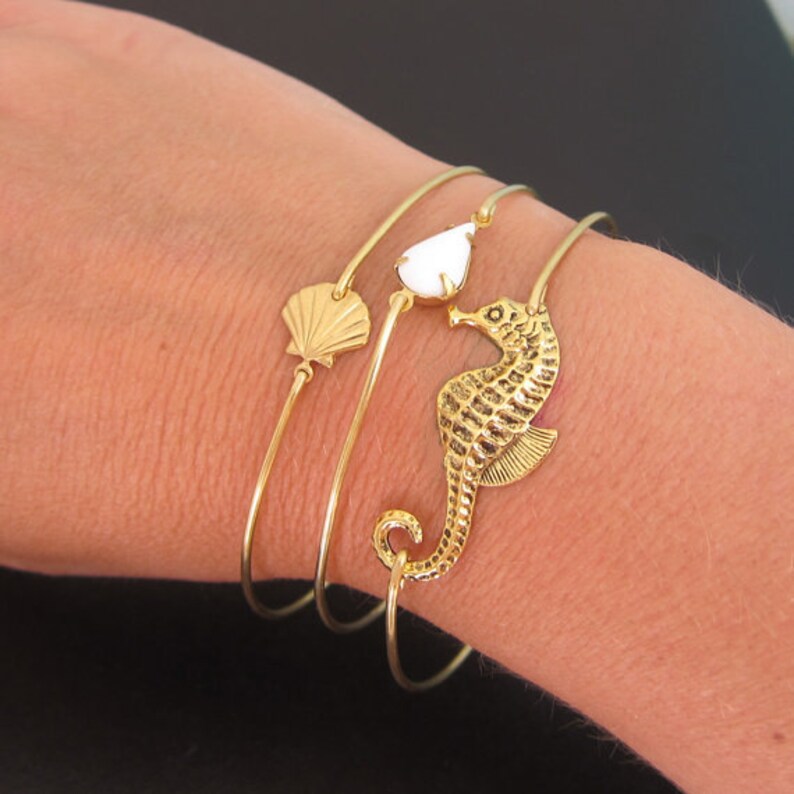 Pode incluir: Pulseiras de ouro com um charme de concha, um charme de l&aacute;grima branca e um charme de cavalo-marinho.