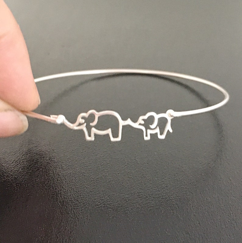 Puede incluir: Un brazalete de plata con dos siluetas de elefantes, una m&aacute;s grande que la otra, conectadas por sus trompas.