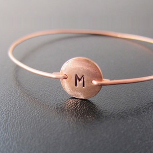 K&ouml;nnte beinhalten: Ein kupferfarbenes Armreif-Armband mit einem kreisf&ouml;rmigen Anh&auml;nger. Der Anh&auml;nger zeigt den Buchstaben "M" in der Mitte. Das Armband ist ein schlichtes, minimalistisches Design f&uuml;r den t&auml;glichen Gebrauch.