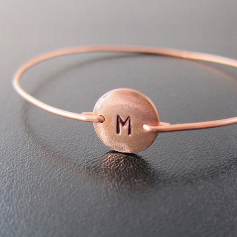 K&ouml;nnte beinhalten: Ein kupferfarbenes Armreif-Armband mit einem kreisf&ouml;rmigen Anh&auml;nger. Der Anh&auml;nger zeigt den Buchstaben "M" in der Mitte. Das Armband ist ein schlichtes, minimalistisches Design f&uuml;r den t&auml;glichen Gebrauch.