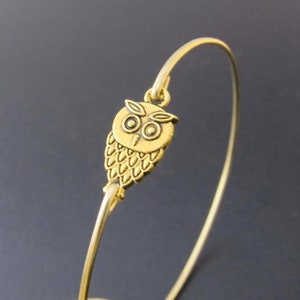 May include: A gold-colored bangle bracelet featuring a detailed owl charm. The owl charm is intricately designed with textured feathers and large, round eyes. The bracelet has a simple, curved design, and the owl charm is attached at the top.
