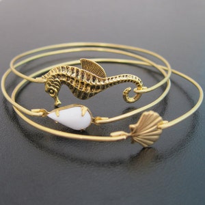 Pode incluir: Tr&ecirc;s pulseiras de bracelete douradas com um amuleto de cavalo-marinho, um amuleto de l&aacute;grima branca e um amuleto de concha.