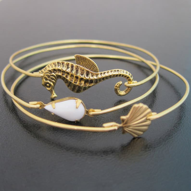 Pode incluir: Tr&ecirc;s pulseiras de bracelete douradas com um amuleto de cavalo-marinho, um amuleto de l&aacute;grima branca e um amuleto de concha.