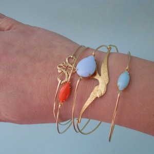 Puede incluir: Cuatro pulseras de brazalete de oro con diferentes encantos. Una pulsera tiene una piedra azul en forma de l&aacute;grima, otra tiene un peque&ntilde;o encanto de p&aacute;jaro, otra tiene una piedra ovalada azul claro y la &uacute;ltima tiene una piedra ovalada de color coral.