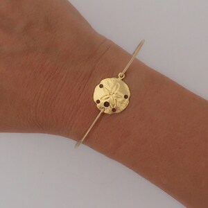 May include: A gold bangle bracelet with a sand dollar charm. The charm is detailed with a five-pointed star in the center.
