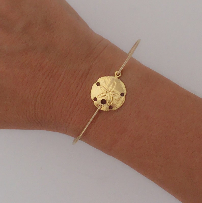 May include: A gold bangle bracelet with a sand dollar charm. The charm is detailed with a five-pointed star in the center.