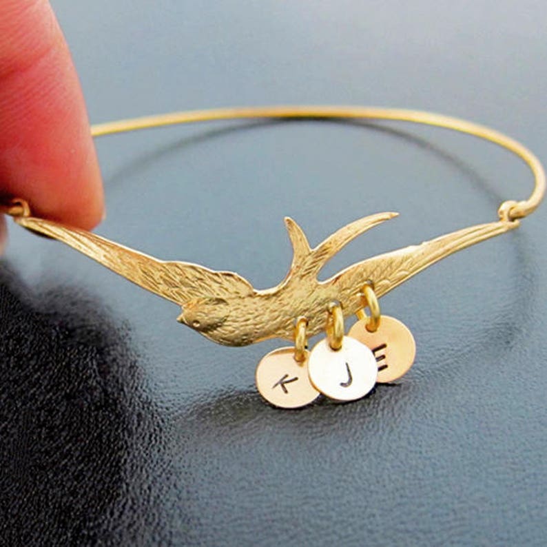 Peut inclure: Un bracelet jonc dor&eacute; avec un motif d'oiseau d&eacute;taill&eacute;. L'oiseau est en vol, avec trois petits charms ronds suspendus en dessous, chacun grav&eacute; d'une lettre. Le bracelet est tenu par un doigt sur un fond sombre.