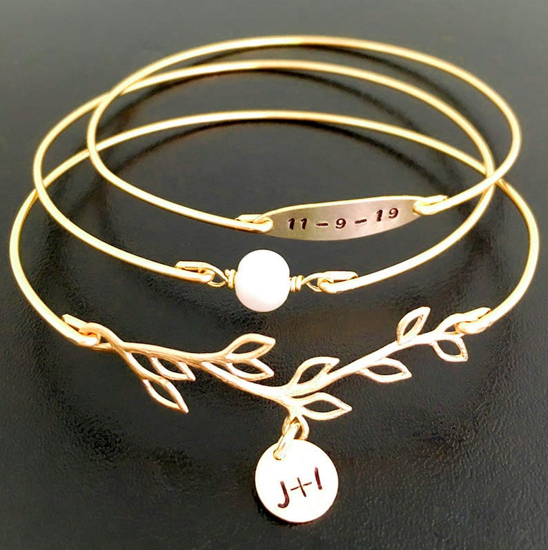 K&ouml;nnte beinhalten: Ein Set aus drei goldenen Armreifen mit einem zarten Zweig-Anh&auml;nger und einem kleinen runden Anh&auml;nger mit den Initialen "J+I". Der mittlere Armreif hat eine kleine wei&szlig;e Perle und ein rechteckiges Schild mit der eingravierten Jahreszahl "11-9-19".