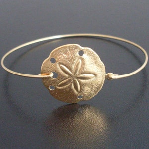 May include: A gold-toned bangle bracelet with a sand dollar charm. The charm has a five-pointed star design and is slightly textured.