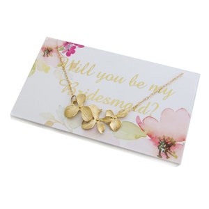 May include: A gold necklace with a floral design and a card that says "Will you be my bridesmaid?" The card has a watercolor floral background.