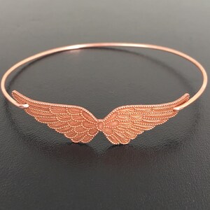 K&ouml;nnte beinhalten: Ein ros&eacute;goldenes Armkettchen mit einem filigranen, detaillierten Engelsfl&uuml;gel-Design.