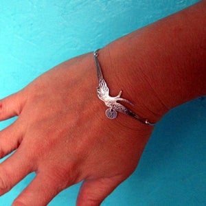 Peut inclure: Bracelet argent&eacute; orn&eacute; d'une hirondelle aux ailes d&eacute;ploy&eacute;es. Un petit charme rond avec la lettre "M" est suspendu en dessous. Le bracelet est port&eacute; sur un poignet, sur fond turquoise.