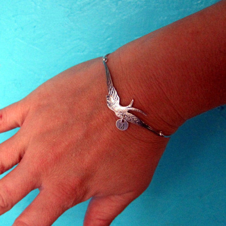 Peut inclure: Bracelet argent&eacute; orn&eacute; d'une hirondelle aux ailes d&eacute;ploy&eacute;es. Un petit charme rond avec la lettre "M" est suspendu en dessous. Le bracelet est port&eacute; sur un poignet, sur fond turquoise.