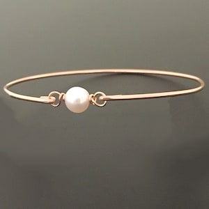 May include: A delicate rose gold bangle bracelet with a single white pearl charm.