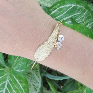 May include: A gold bangle bracelet with a large gold wing charm and small charms with letters. The bracelet is on a person's wrist.