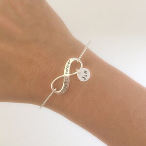 May include: Silver infinity bangle bracelet with the words "together forever" engraved on the infinity symbol. A small charm with the initials "J & B" hangs from the bangle.