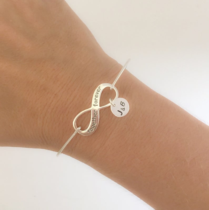 May include: Silver infinity bangle bracelet with the words "together forever" engraved on the infinity symbol. A small charm with the initials "J & B" hangs from the bangle.