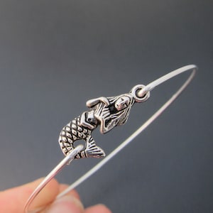 Puede incluir: Una pulsera de plata con un dije de sirena. La sirena est&aacute; mirando hacia la derecha y tiene un dise&ntilde;o detallado.