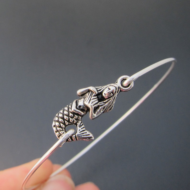 Puede incluir: Una pulsera de plata con un dije de sirena. La sirena est&aacute; mirando hacia la derecha y tiene un dise&ntilde;o detallado.