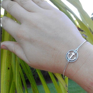 K&ouml;nnte beinhalten: Ein silbernes Armreif-Armband mit einem Anker-Anh&auml;nger. Das Armband befindet sich am Handgelenk.