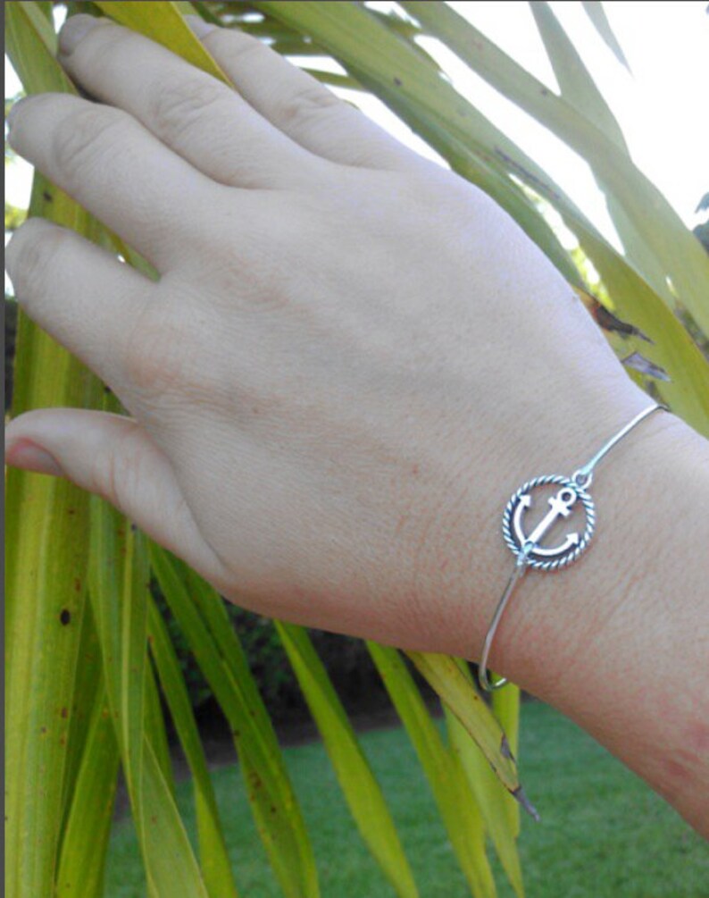 K&ouml;nnte beinhalten: Ein silbernes Armreif-Armband mit einem Anker-Anh&auml;nger. Das Armband befindet sich am Handgelenk.