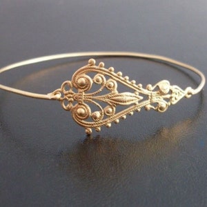 May include: A gold-colored, filigree-style bracelet. The bracelet features an ornate, symmetrical design in the center, with delicate swirls, dots, and a central leaf-like element. The thin band of the bracelet is circular.