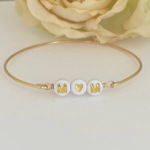 May include: A gold bangle bracelet with white beads that spell out "MOM" with a gold heart in between the letters.