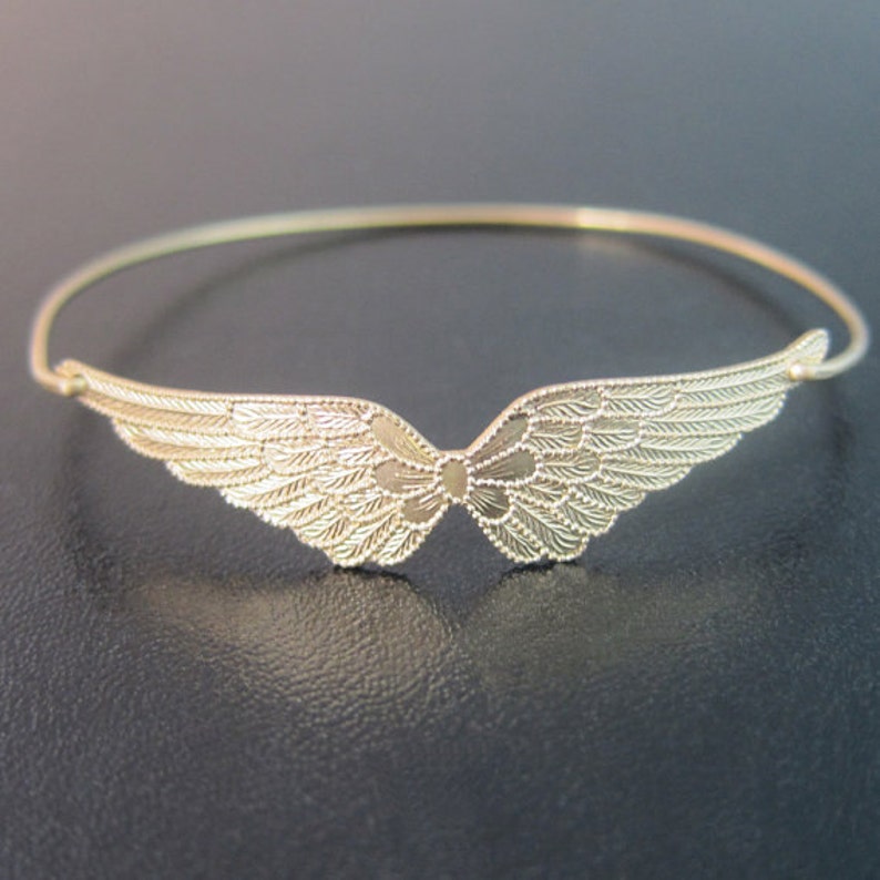 K&ouml;nnte beinhalten: Ein goldfarbenes Armreif-Armband mit einem filigranen, detaillierten Design von Engelsfl&uuml;geln.
