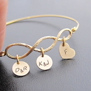 May include: Gold infinity bangle bracelet with three charms. The charms are round with the initials "D&R", "K&J" and a heart charm with the initial "F".