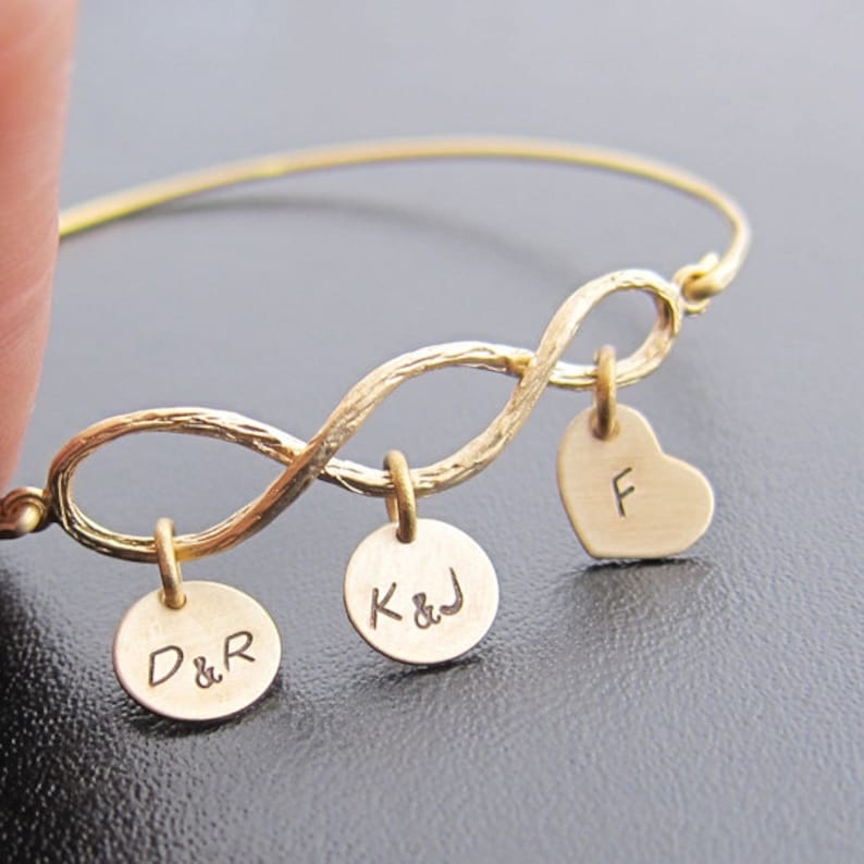 May include: Gold infinity bangle bracelet with three charms. The charms are round with the initials "D&R", "K&J" and a heart charm with the initial "F".