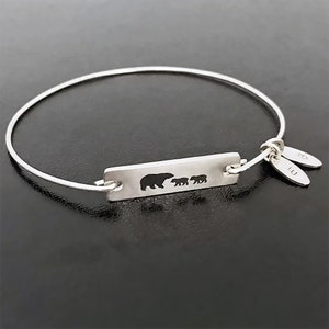 May include: A silver bangle bracelet with a rectangular charm featuring a silhouette of a mother bear and two cubs. The charm is engraved with the letters "E" and "B".
