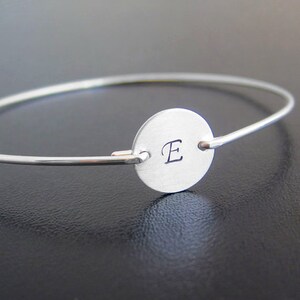 May include: A silver bangle bracelet with a round charm featuring the letter "E" engraved on it.