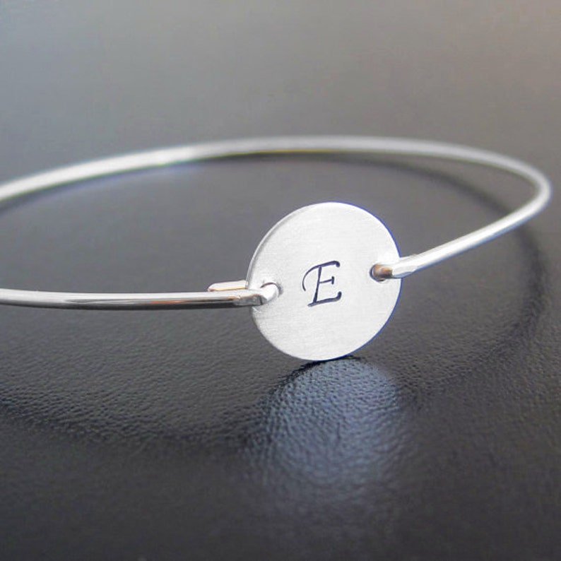 May include: A silver bangle bracelet with a round charm featuring the letter "E" engraved on it.