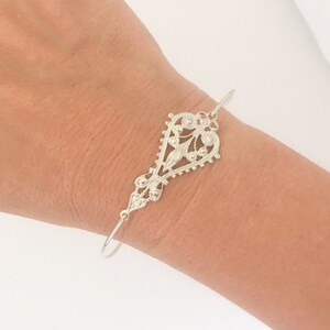 May include: A silver filigree bracelet with a heart-shaped design. The bracelet features intricate openwork patterns and a delicate, ornate appearance. The bracelet is worn on a wrist.