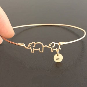 May include: Gold bangle bracelet with two elephant charms and a small round charm with the letter 'M' engraved on it.