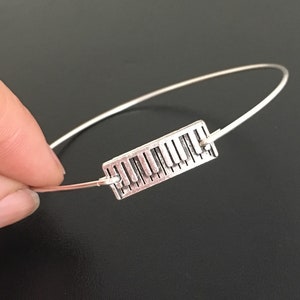 May include: A silver bracelet with a piano key charm. The charm is a rectangular shape with a detailed design of piano keys.