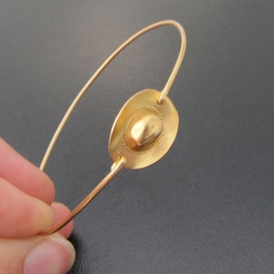 May include: A gold-toned bracelet with a circular charm featuring a raised, textured design. The charm is attached to a thin, wire-like band.