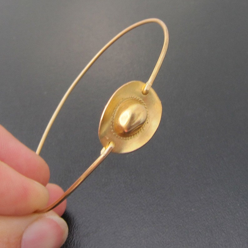 May include: A gold-toned bracelet with a circular charm featuring a raised, textured design. The charm is attached to a thin, wire-like band.