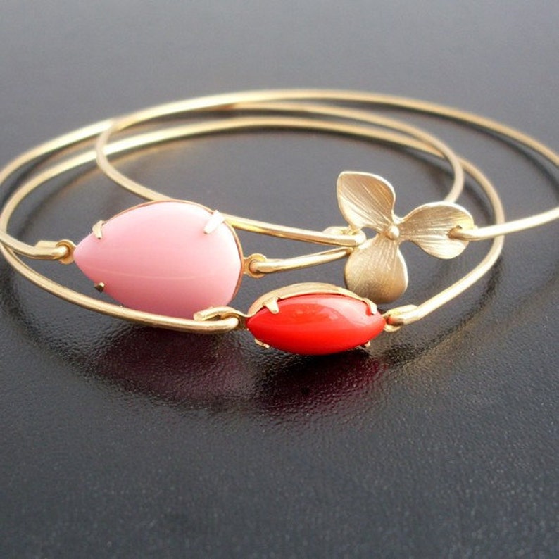 Op de afbeelding: Drie gouden armbandjes met roze en rode glazen accenten. Het middelste armbandje heeft een gouden bloemenbedel.