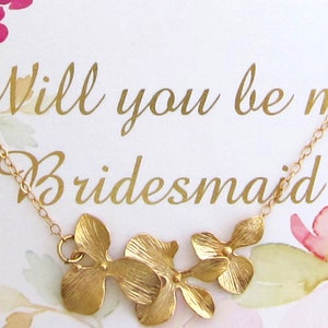 May include: A gold necklace with a floral design and the text "Will you be my bridesmaid?" on a white card with watercolor flowers.