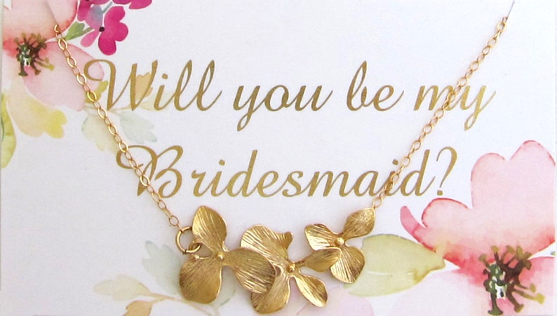 May include: A gold necklace with a floral design and the text "Will you be my bridesmaid?" on a white card with watercolor flowers.
