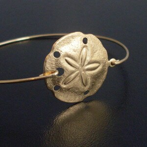 May include: A gold-toned bangle bracelet with a sand dollar charm. The charm is detailed with a five-pointed star and textured surface.